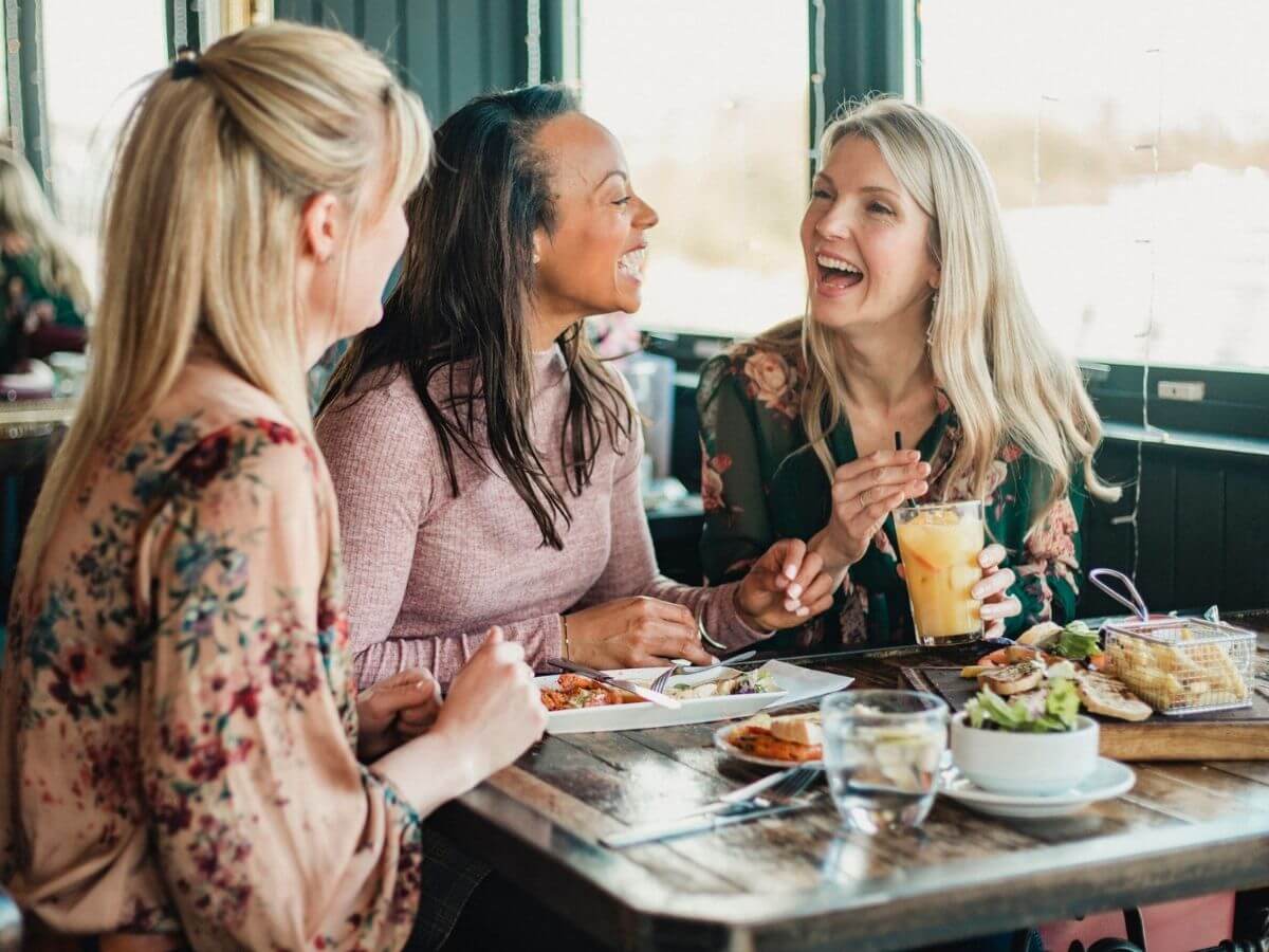A group of three women laugh together at a restaurant over brunch.