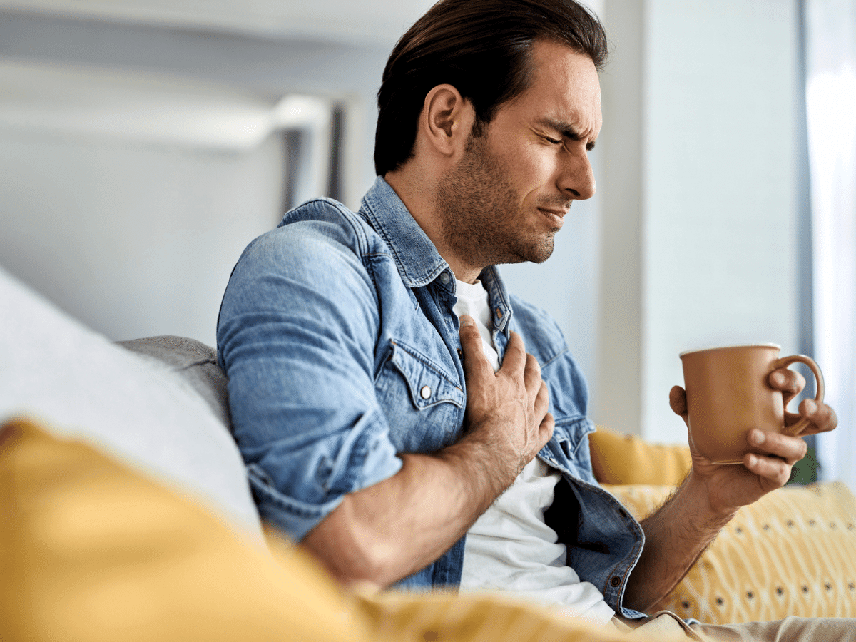 Young man at home sitting on couch drinking tea, holds chest in pain